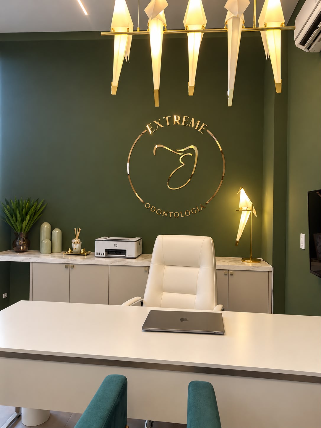 Parede verde com o logo dourado da Extreme Odontologia, luminárias em forma de pássaro e mesa de atendimento clara